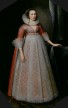 A standing young woman resting her hand on a table and wearing a floor-length gown covered in delicate white lace