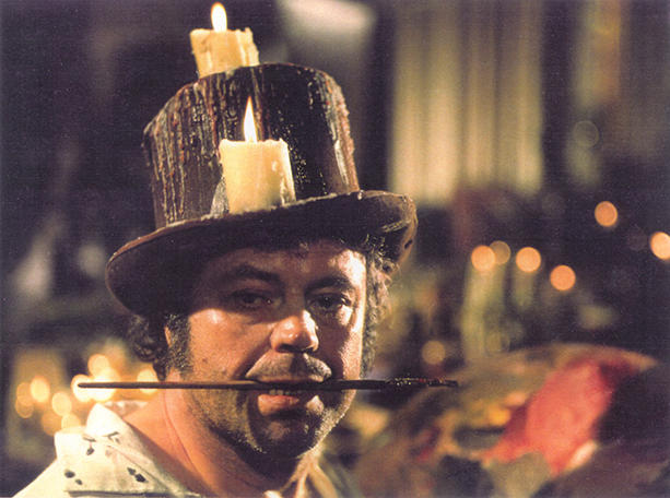 A man with an old top hat one with candles on it holding a paint brush in his mouth