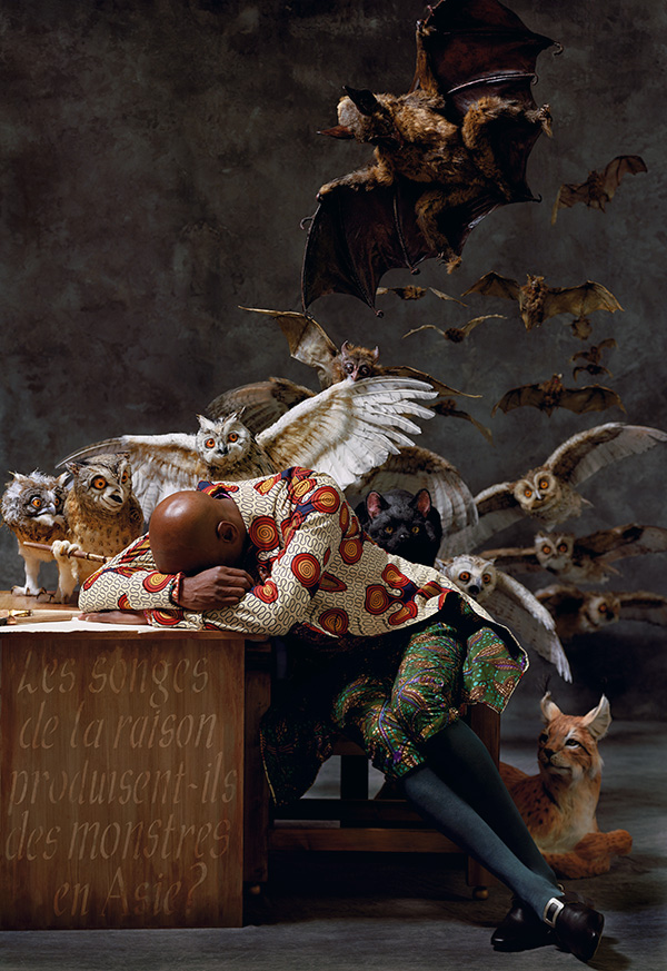 A color photograph of a seated man with his head rested on his arms on a desk, behind him are owls, bats and a lynx