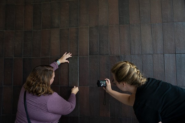Two women standing in front of a brown tiled wall with their backs turned to the viewer, the woman on the left holds up a brown tile against the wall and the woman on the right takes a photograph of this action